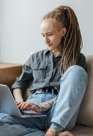 Image of a concentrated young girl with dreadlocks and piercing indoors using laptop computer.