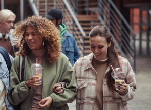 Group of cheerful intercultural teenage girls with soda having chat while walking down street in front of their boyfriends outdoors